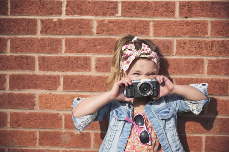 A little girl prepares to take a photo next to a brick wall.